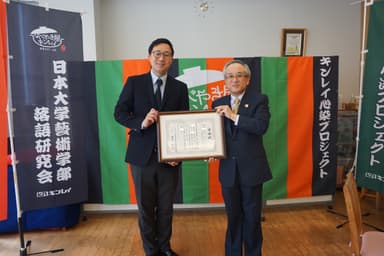 授与式の様子　右：湯下 廣一会長　左：商品企画部マネジャー・福田 暢雄