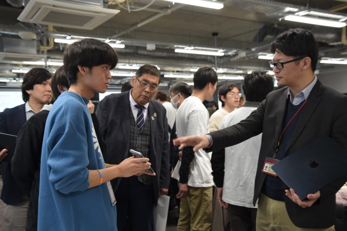 学生の説明を聞く小原学長や情報工学部の鈴木秀和准教授(右)