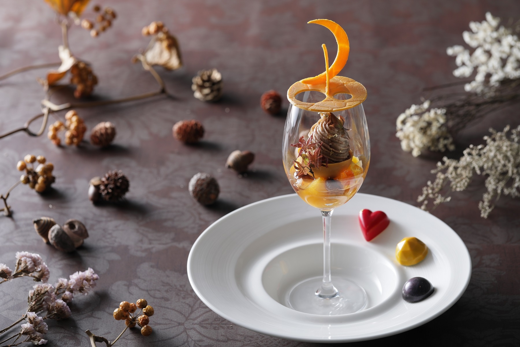 【Dessert】 秋の夜長のお月見から着想したチョコレートのアントルメが印象的なアートパフェ。 ラム酒の効いたラムレーズンアイスのまわりを マロンクリームの絞りでモンブラン仕立てに。 下層はトパーズをイメージした黄色いチョコレートの中にコクのあるチーズケーキをin。 柑橘系のお酒で香付けしたジュエルイメージの琥珀糖や栗の渋皮煮、 フルーツのオレンジがアクセントになっています。 また、目が奪われるような炎のような色、情熱を連想させることからガナッシュにパッションフルーツを使用したファイヤーオパール、友情という石言葉から相性の良いキャラメルとオレンジをチョイスしたトパーズ、タンザニアの夜空をイメージし香り高いキリマンジャロコーヒーをガナッシュに使用したタンザナイト、3種のボンボンショコラが主役のアートパフェを引き立てます。