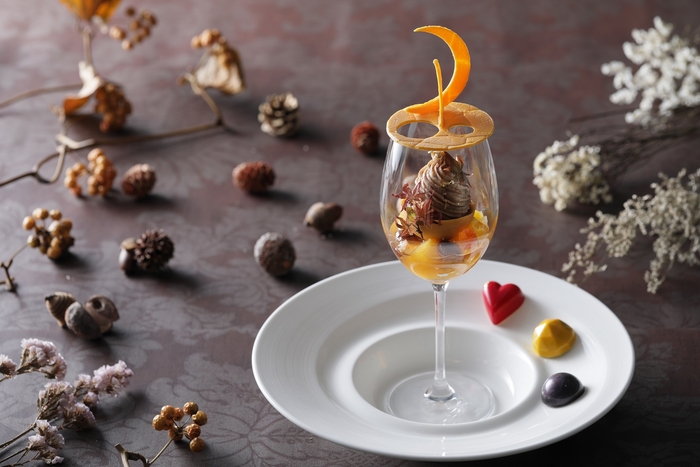 【Dessert】 秋の夜長のお月見から着想したチョコレートのアントルメが印象的なアートパフェ。 ラム酒の効いたラムレーズンアイスのまわりを マロンクリームの絞りでモンブラン仕立てに。 下層はトパーズをイメージした黄色いチョコレートの中にコクのあるチーズケーキをin。 柑橘系のお酒で香付けしたジュエルイメージの琥珀糖や栗の渋皮煮、 フルーツのオレンジがアクセントになっています。 また、目が奪われるような炎のような色、情熱を連想させることからガナッシュにパッションフルーツを使用したファイヤーオパール、友情という石言葉から相性の良いキャラメルとオレンジをチョイスしたトパーズ、タンザニアの夜空をイメージし香り高いキリマンジャロコーヒーをガナッシュに使用したタンザナイト、3種のボンボンショコラが主役のアートパフェを引き立てます。