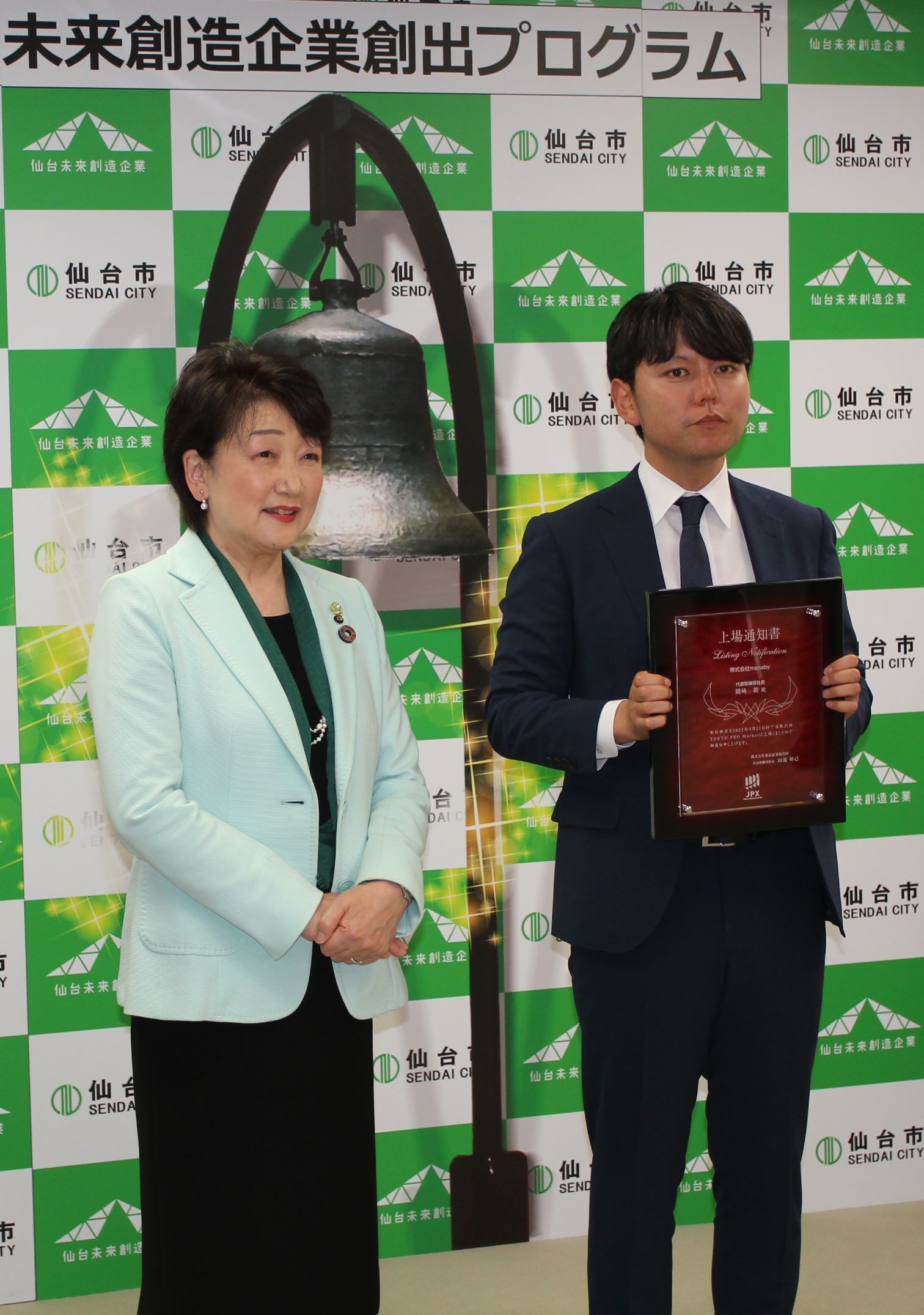 鐘をプリントしたパネルの前で、上場を祝う岡崎社長(右)と郡市長